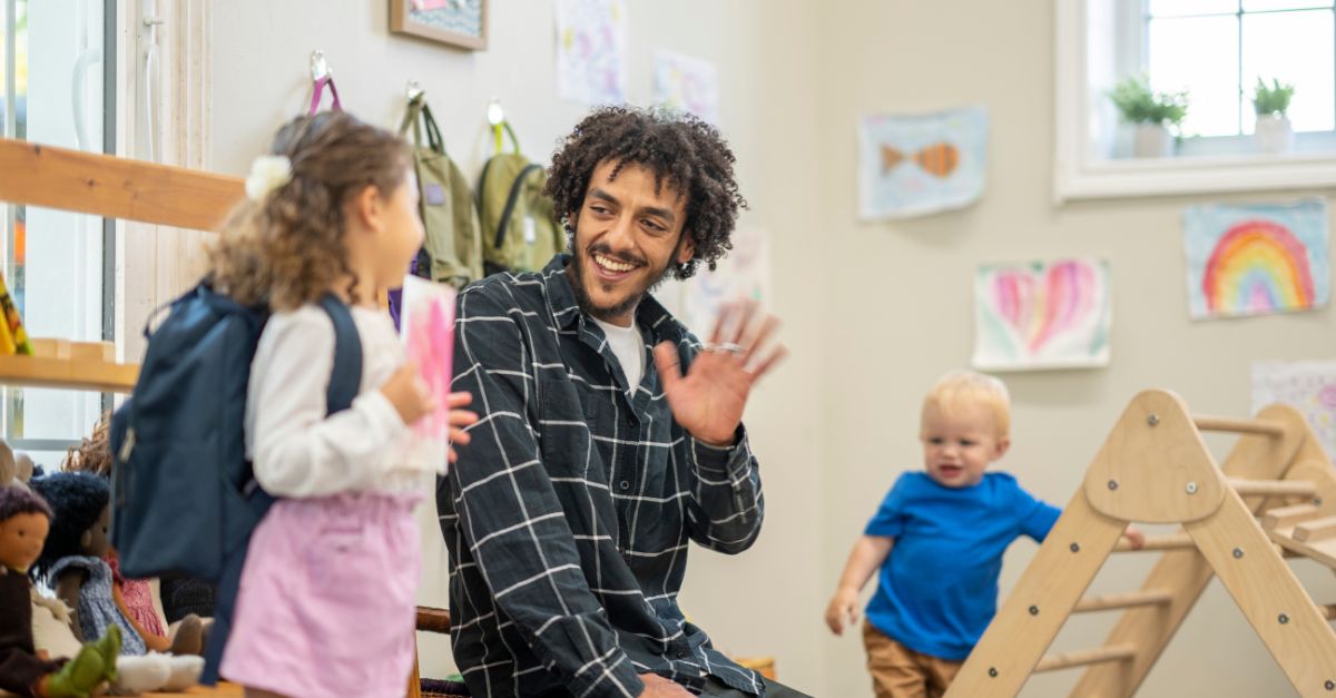 Preschool teacher waving and welcoming young girl into the classroom on her first day of school