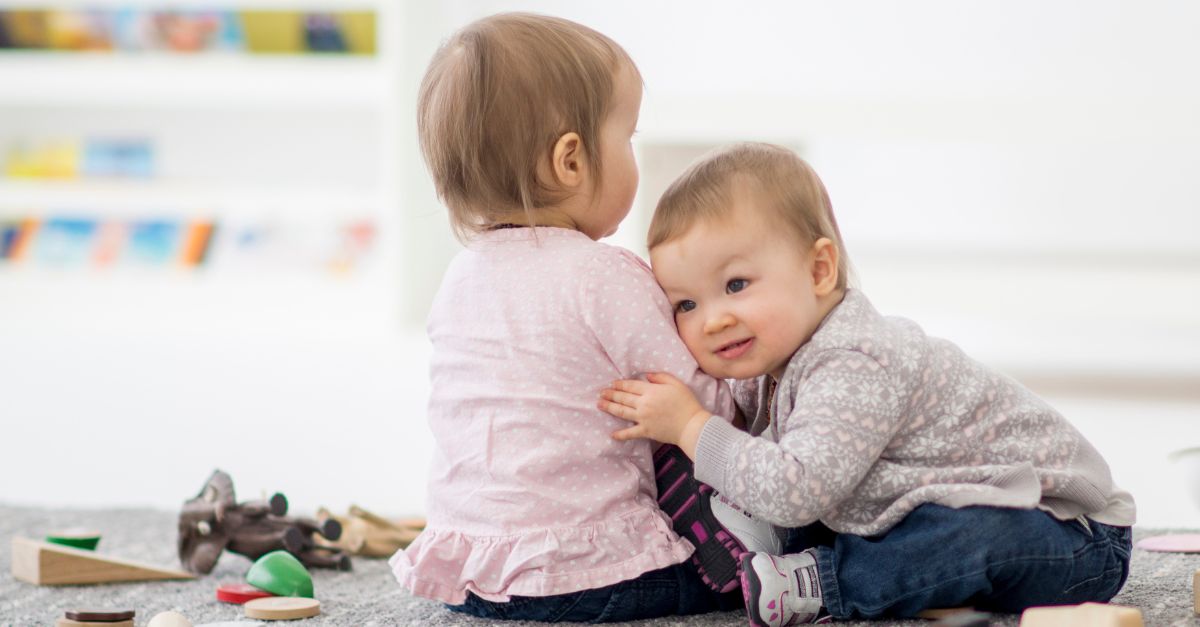 One infant leaning on and hugging another as they play in their infant classroom