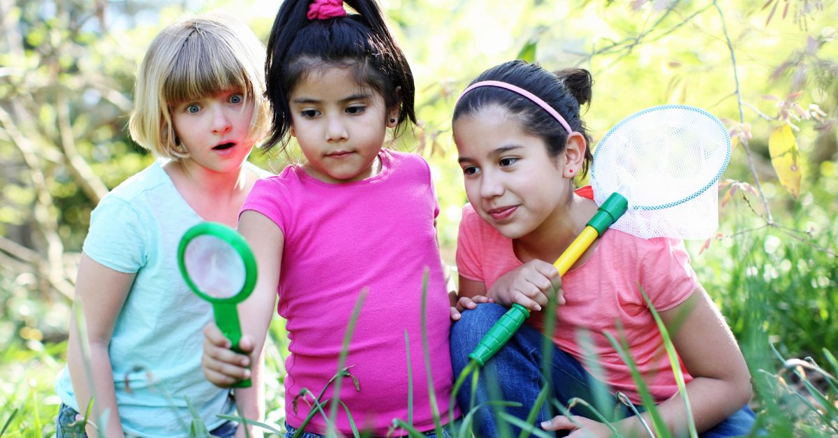 Three preschool girls exploring outside with magnifying glasses and bug nets