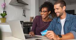 Two parents sitting behind a computer screen, smiling, with a credit card in hand as they pay their childcare payments online from their home. 