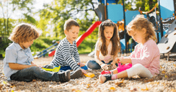 Four preschool children sitting on the ground and exploring rocks and natural materials with small shovels and cups in the playground