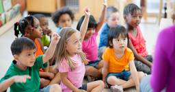 A group of kindergarten aged children sitting on the floor in front of their teacher, with their hands raised for circle time. 
