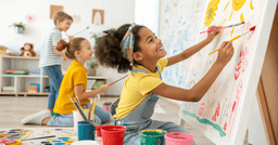 Preschool girl in the foreground smiling and painting flowers and sun on a large canvas, with two other preschoolers painting in the background