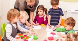 A group of preschool aged children playing with crayons, paper and playdough at the art table. A teacher in the background helping children with the materials on the table. 