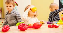Three toddlers playing make-believe with with pretend cups and food at a table in their childcare classroom. 