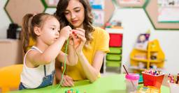 A preschool teacher demonstrating scaffolding techniques as she supports a student in placing beads on a string. 