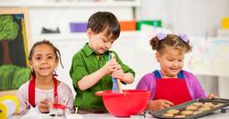 Three kindergarten children in aprons, whisking baking items together and baking cookies. 