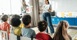 Two early childhood educators showing learning materials to a group of preschoolers sitting on chairs