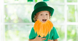 A preschool aged boy playing dress up with a green shirt, green hat and pretend orange beard for St.Patricks day. 