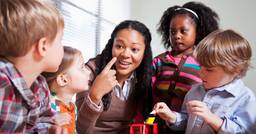An early childhood educator sitting at a table with four children as she points to her eye and engages in conversation. 