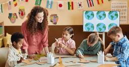 Four preschoolers sitting at a table decorating cardboard flowers and trees. Preschool teacher standing at the table. 