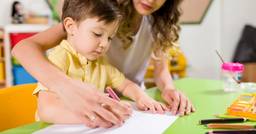 A preschool teacher supporting the hand of a young boy as he practices tracing the alphabet at daycare. 