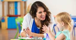 An early childhood educator sitting at a table, taking notes and smiling at a preschool who is practicing clapping. 