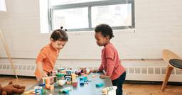 Two preschoolers engaged in parallel play with blocks at a table. One preschooler is observing how the other is manipulating the blocks. 