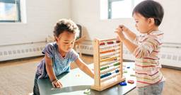 Two preschool aged children playing with and sharing cognitive materials across from each other at a table in their classroom. 