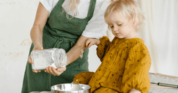 Preschool teacher helping preschooler pour an ingredient into a bowl during cooking lesson.