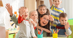 Early childhood educator smiling in the classroom with 5 students smiling and waving at the camera