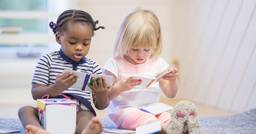 Two preschoolers sitting side by side  on the carpet in their classroom, reading books separately. 