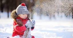 A preschool aged boy playing outside in the winter, blowing snow from his mittens. 