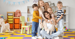 A home childcare provider sitting in their classroom with 5 children standing beside them and smiling at the camera. 