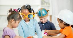 Four preschool aged children engaged in dramatic play in the preschool classroom, wearing large glasses and hats on their heads as they share materials. 