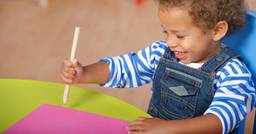 An older infant sitting at a table practicing fine motor skills by holding a large crayon and moving it across a piece of paper 