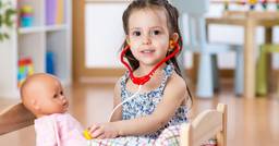 Little girl holding a baby doll and wearing a plastic stethoscope while playing doctor at preschool. 