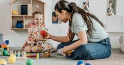 A preschool demonstrates scaffolding techniques as she sits on the floor beside a child and hands her a red ball to encourage the advancement of her play