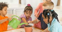 A preschool aged boy using a plastic watering can to water seeds in a pot, with four other children around him exploring the seeds and dirt. 
