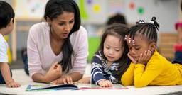 Early childhood educator and two preschoolers reading a book on the carpet