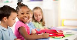 A preschool child smiling and drawing with crayons, with three other children around her in the preschool classroom. 