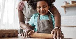 Preschool teacher helping preschooler roll dough during cooking lesson.
