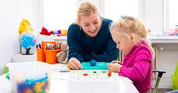 Preschool teacher and child sitting at table, playing with play dough 
