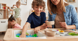 Preschool boy and preschool teacher in the foreground experimenting with a spring sensory bin. Two other preschoolers playing in the background. 