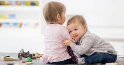 One infant leaning on and hugging another as they play in their infant classroom