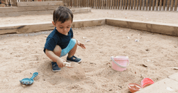 Toddler squatting down in the sandbox and scooping sand with a shovel