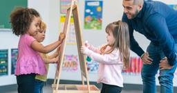 Three preschool aged children playing at an art easel, two on one side and one on the other, as they draw with markers. A teacher in the background observing and smiling at their work. 