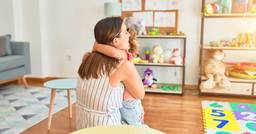 An early childhood educator and toddler hugging in the preschool classroom on a sunny day. 