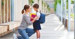 Mom on her knees in front of her kindergarten aged daughter with her arms around her and encouraging her as she goes into her first day of school. 
