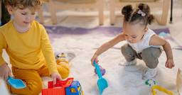 Two preschoolers playing side by side in the sandbox while they dig with small shovels and toy trucks. 