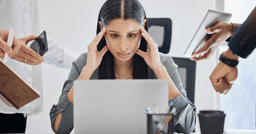 A home childcare provider sitting at their computer with their hands on their temples showing signs of overwhelm as multiple staff show hand them work and questions for review.